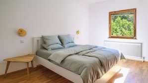 A bedroom with a wooden floor, a bed with a grey bedspread, a small wooden table, and a window with a view of greenery.