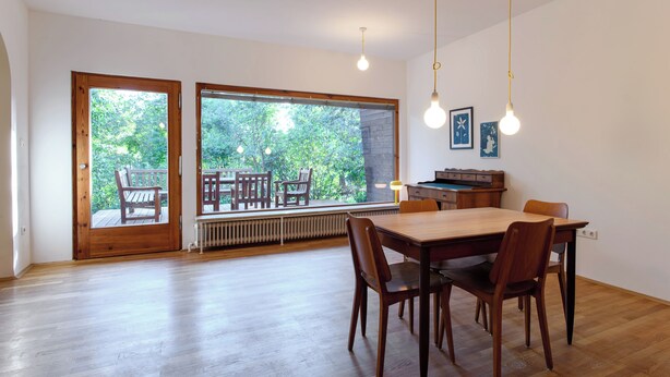 A spacious dining area with a wooden table and chairs, large windows with a view of the outdoors, and hanging pendant lights.