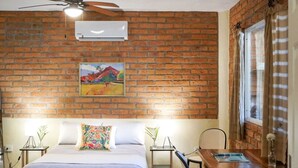 A hotel room with a brick wall, a bed with a colorful pillow, a ceiling fan, an air conditioner, a window with curtains, and a small table with a chair. - Casa Redonda 🌼 Casita 1 🌞SayulitaWifi 💛 Central (Sayulita)