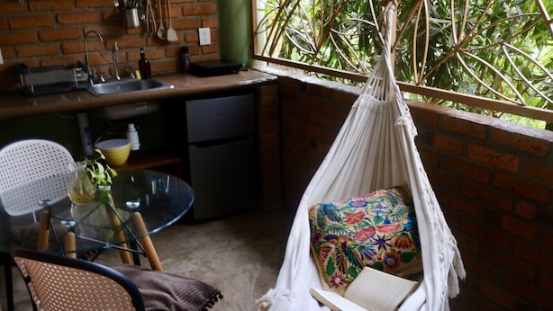 A hammock chair with a colorful pillow, a glass table with chairs, and a kitchen area with a sink and utensils. - Casa Redonda 🌼 Casita 1 🌞SayulitaWifi 💛 Central (Sayulita)
