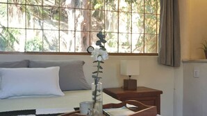 Un dormitorio con cama, mesita de noche con lámpara, bandeja con un florero y ventana con vistas a árboles.
