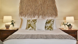 Una cama con cabecera de tejido, ropa de cama blanca y almohadones decorativos. Hay dos mesitas de noche con lámparas y jarrones.