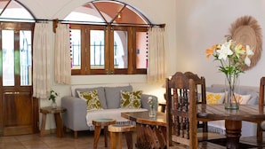Een gezellige woonkamer met een bank, een houten eettafel en stoelen. Er zijn twee ramen met gordijnen, een klein bijzettafeltje en een decoratief wandobject.
