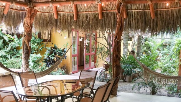 A thatched-roof outdoor area with a hammock, chairs, and potted plants.