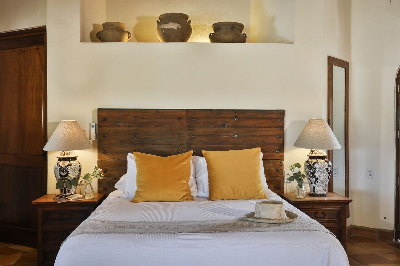 A neatly made bed with two lamps on either side, a wooden headboard, and a shelf above with pottery.