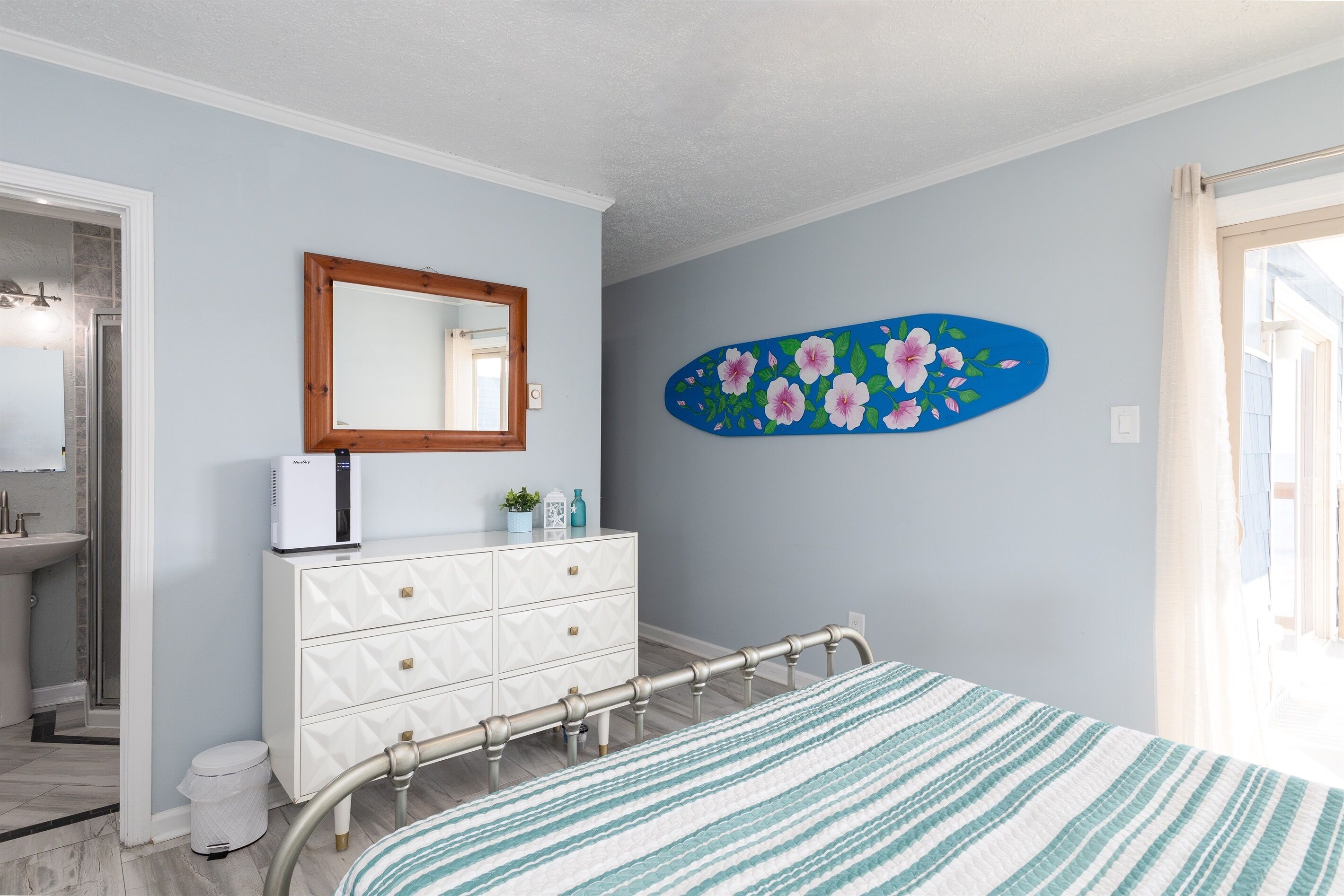 Une chambre à coucher comprenant un lit, une commode, un miroir et un mur orné d’un tableau représentant des motifs floraux.