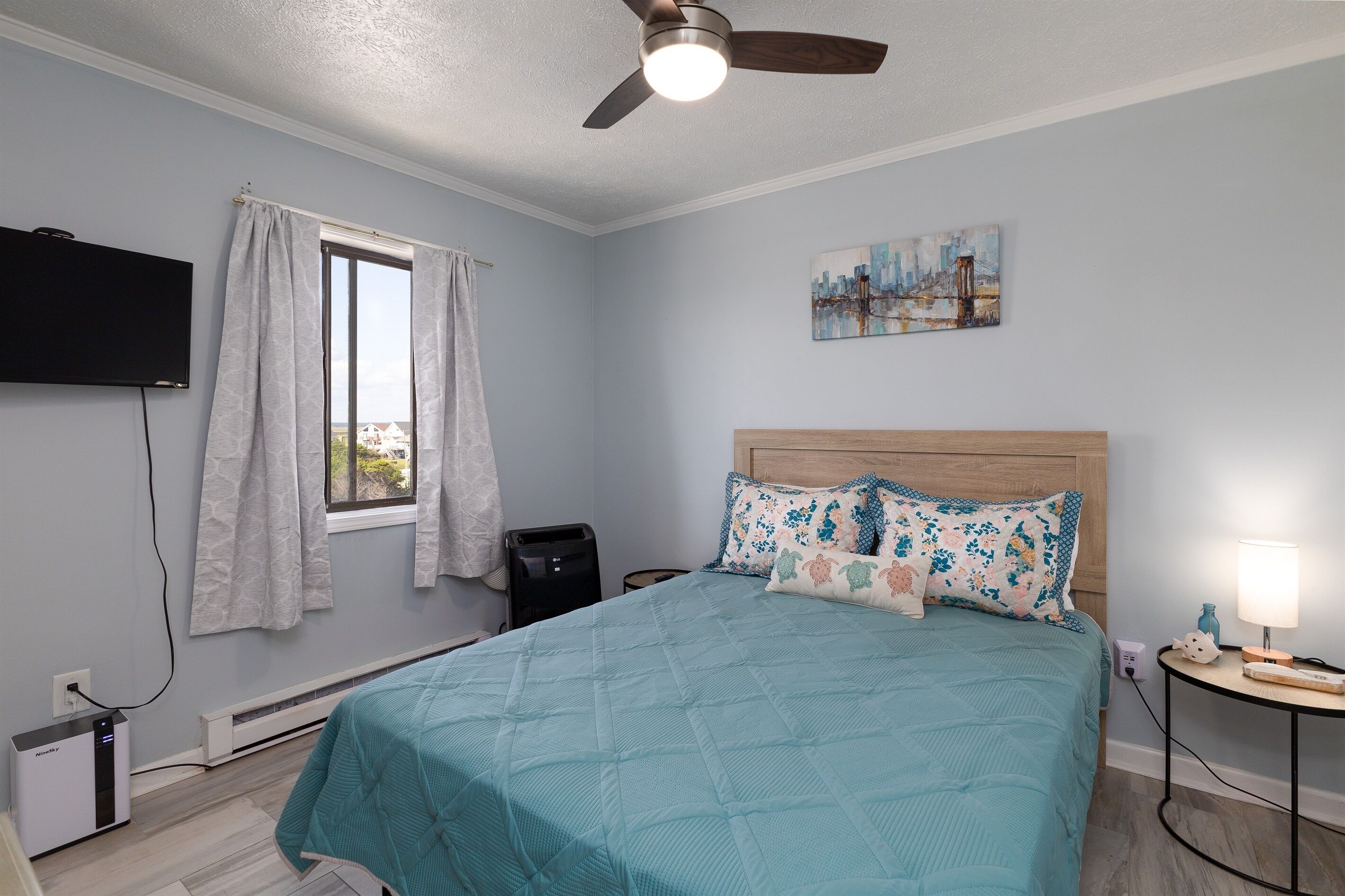 Un dormitorio con una cama, ventilador de techo, ventana con cortinas, calefactor, televisor montado en la pared y un cuadro en la pared.