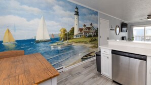 A kitchen with a mural of a lighthouse and sailboats, white cabinetry, and a wooden table.