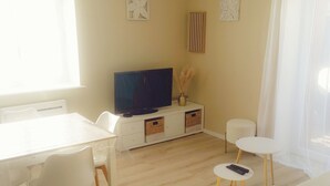 Een moderne, goed verlichte kamer met een flatscreen televisie, een eettafel met stoelen, een klein wit tafeltje met een afstandsbediening, een bed en een raam met dunne gordijnen.