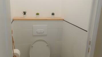 A small, clean bathroom with a toilet, bidet, and two small potted plants on a shelf above the toilet.