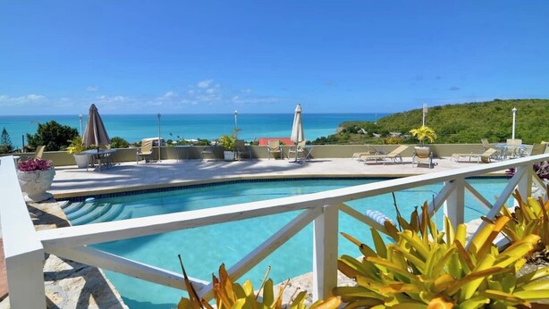 A pool area with lounge chairs and umbrellas, overlooking a clear blue sea and a hilly landscape. - Paradise View - Stunning views near Dickenson Bay Beach in Antigua & Barbuda (Dickenson Bay Beach)