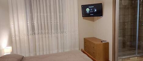 Un dormitorio con cama, televisor montado en la pared, mesita de noche de madera y puerta corrediza de cristal que da a la zona de ducha.