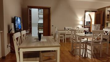 A dining area with a wooden table and white chairs, a television on the left, and a kitchen area visible through an open door.