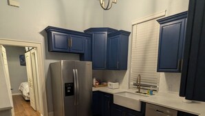 Cozinha com armários azul escuro, uma geladeira de aço inoxidável, bancada branca e piso de madeira.