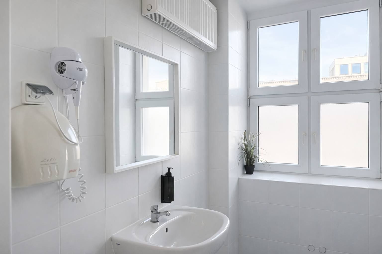 A bathroom with white tiles, a hairdryer, a sink, and a window with a view of buildings.