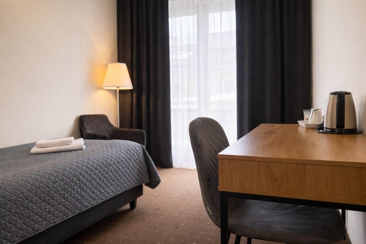 Une chambre d’hôtel avec un lit, un bureau, une chaise, une lampe et une fenêtre avec des rideaux.