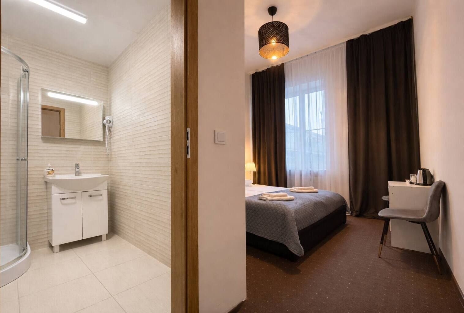 A bathroom with a shower, sink, and mirror, and a bedroom with a bed, chair, and window.