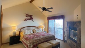 A bedroom with a bed, a ceiling fan, a nightstand, a lamp, a bench, a fireplace, and a window with blinds. - Mountain retreat! With spectacular views! (Burnsville)