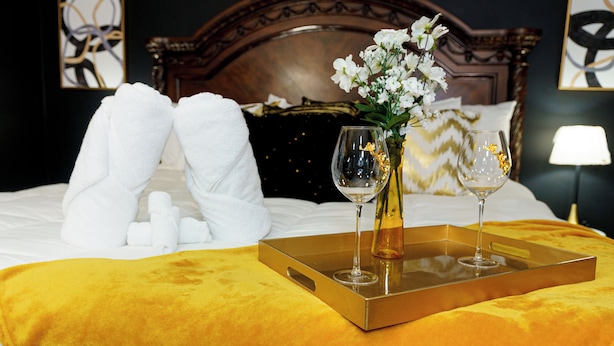 Ein ordentlich bezogenes Bett mit weißen Handtüchern, ein goldener Tablett mit zwei Weingläsern und eine Vase mit weißen Blumen.