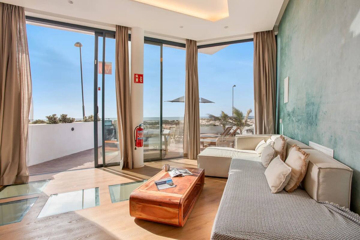 A modern hotel room with a large sliding glass door leading to a balcony, a sofa, a coffee table, and a view of the beach.