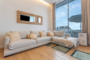 A modern living room with a sectional sofa, a large window with curtains, a mirror, and a small table with a cup. - MAAR Suites Cotillo (La Oliva)