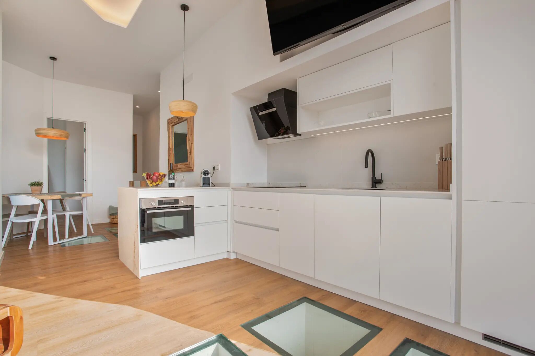 A modern kitchen with white cabinets, a built-in oven, a black faucet, and a flat-screen TV mounted above the counter.