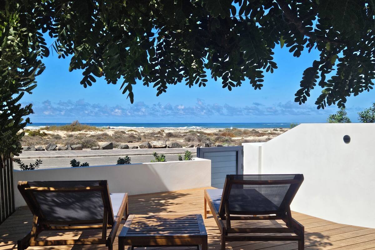 A deck with two wooden chairs and a table, overlooking a beach and ocean.