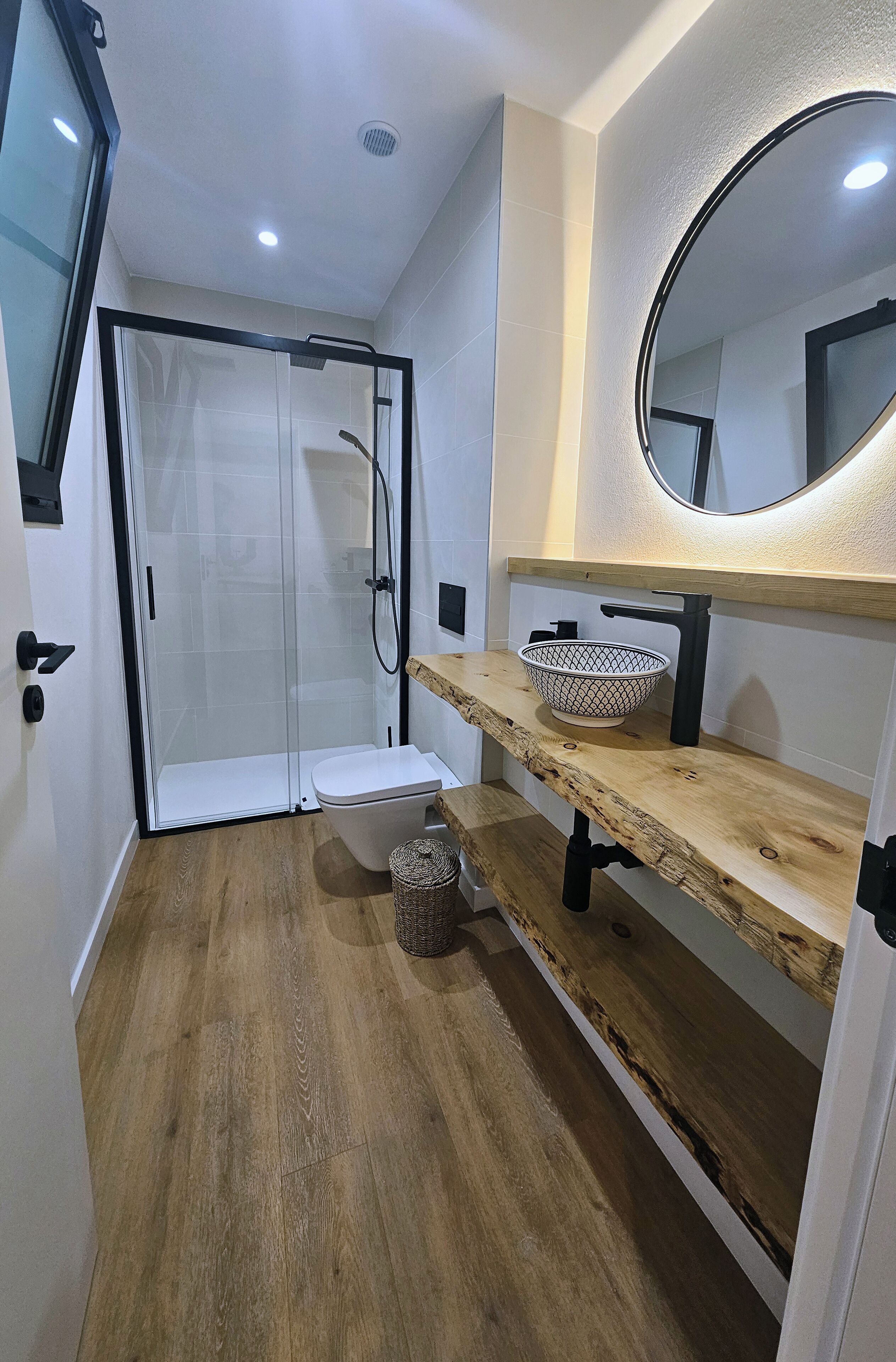 Un baño moderno con cabina de ducha de vidrio, inodoro blanco, tocador de madera con cesta y espejo redondo.