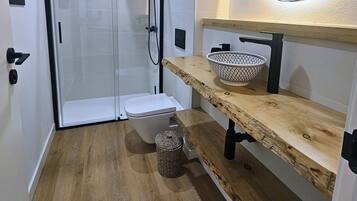 Un baño moderno con cabina de ducha de vidrio, inodoro blanco, tocador de madera con cesta y espejo redondo.