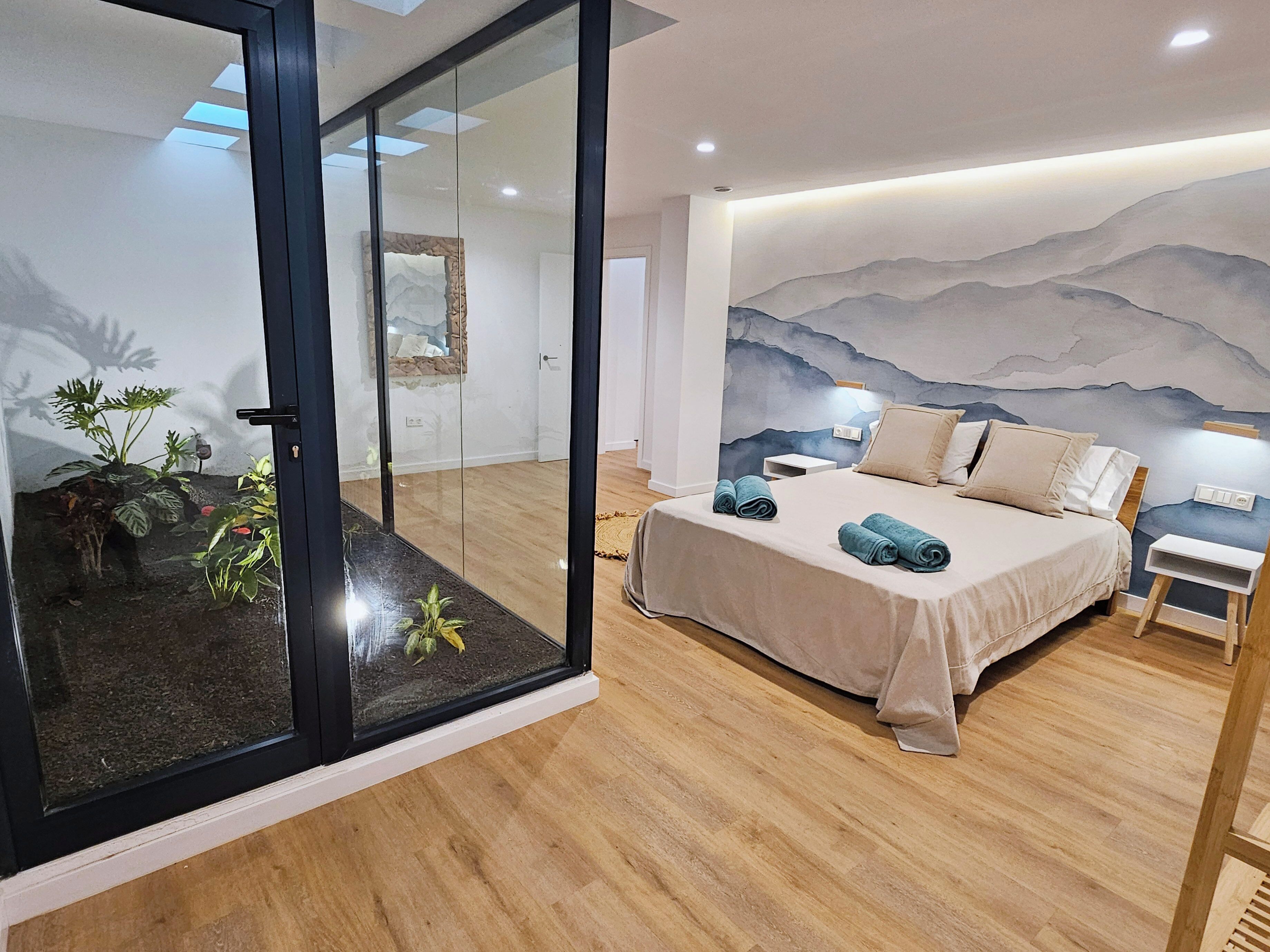 A bedroom with a bed, a nightstand, a wall-mounted lamp, and a glass door leading to a room with plants.