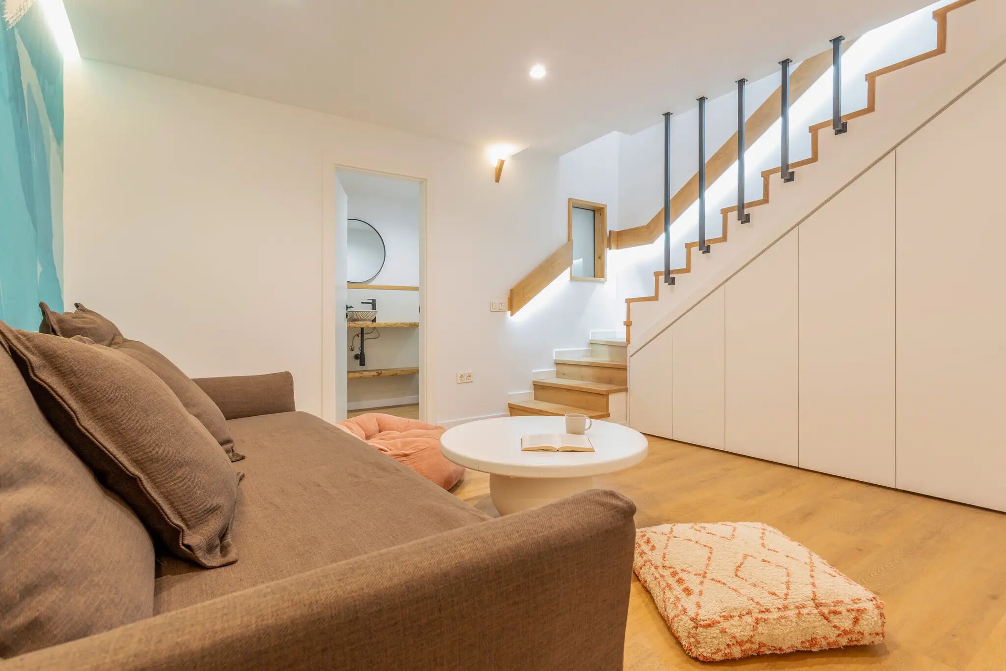 Una sala moderna con un sofá marrón, una mesa redonda de centro, una escalera con escalones de madera y una pared blanca con un espejo.