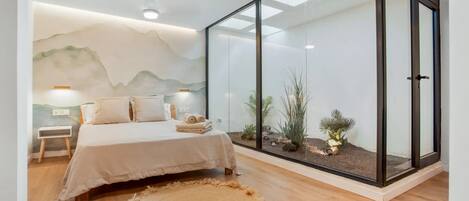 A bedroom with a bed, bedside table, lamp, and a large window with a view of plants.