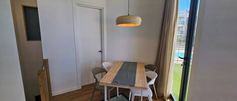 A modern room with a wooden dining table, white chairs, a hanging light fixture, and a view of a swimming pool through a large window.