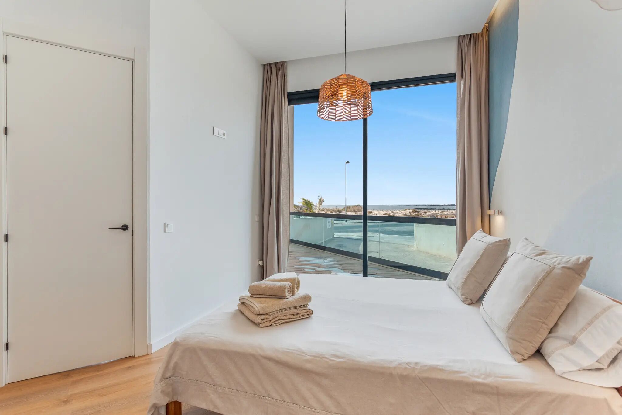 Un dormitorio con cama, lámpara colgante, ventana con vista a una piscina y una playa, y una puerta.