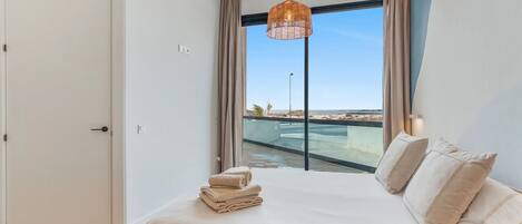Un dormitorio con cama, lámpara colgante, ventana con vista a una piscina y una playa, y una puerta.