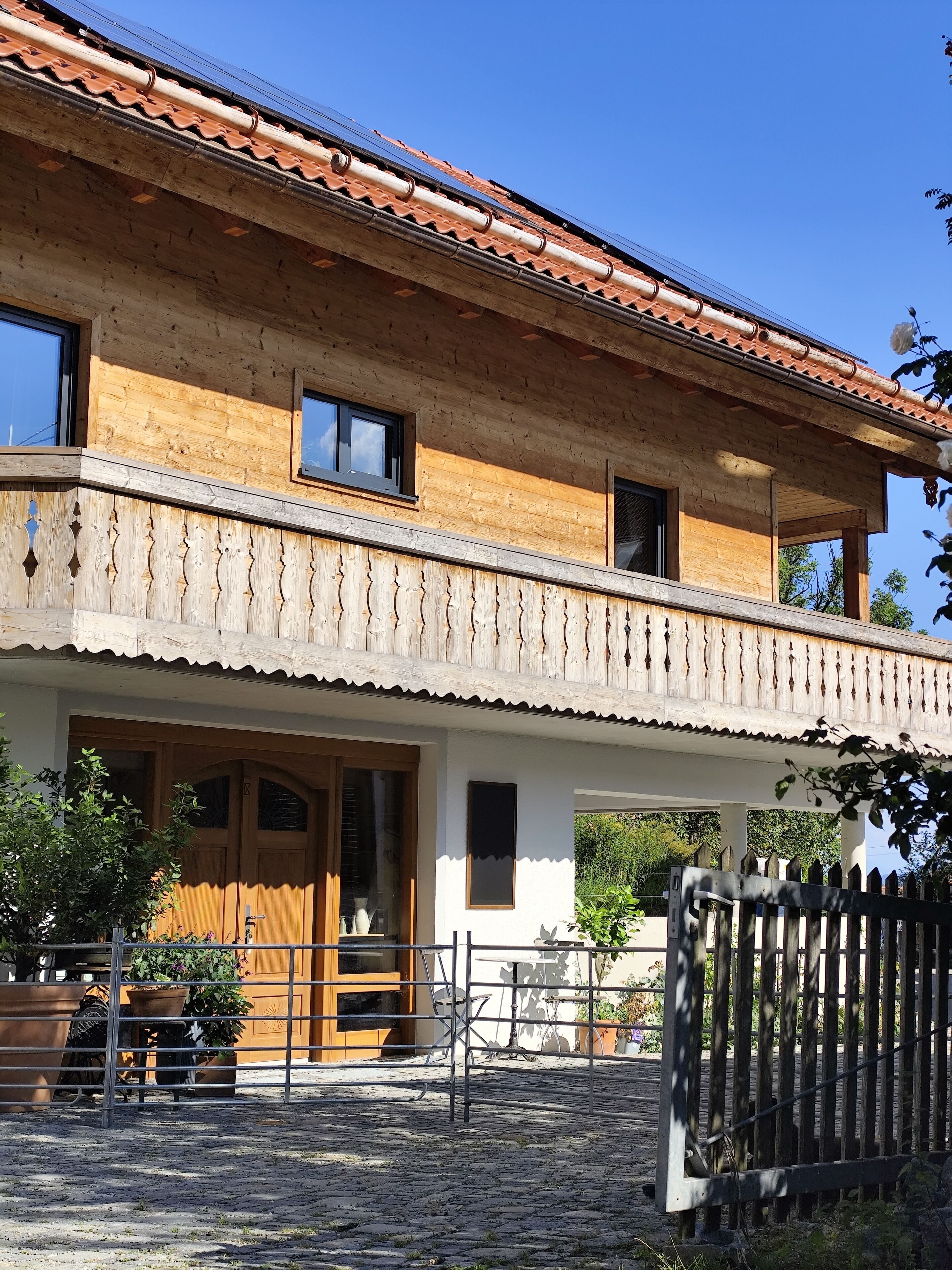 A two-story wooden house with a tiled roof, a balcony with a railing, and a gated entrance.