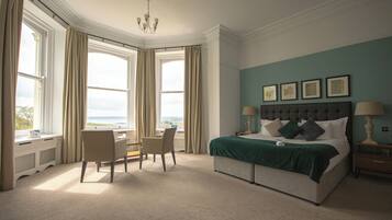 A bedroom with a large bed, a desk, and a chair. There are two windows with curtains, a chandelier, and framed pictures on the wall.