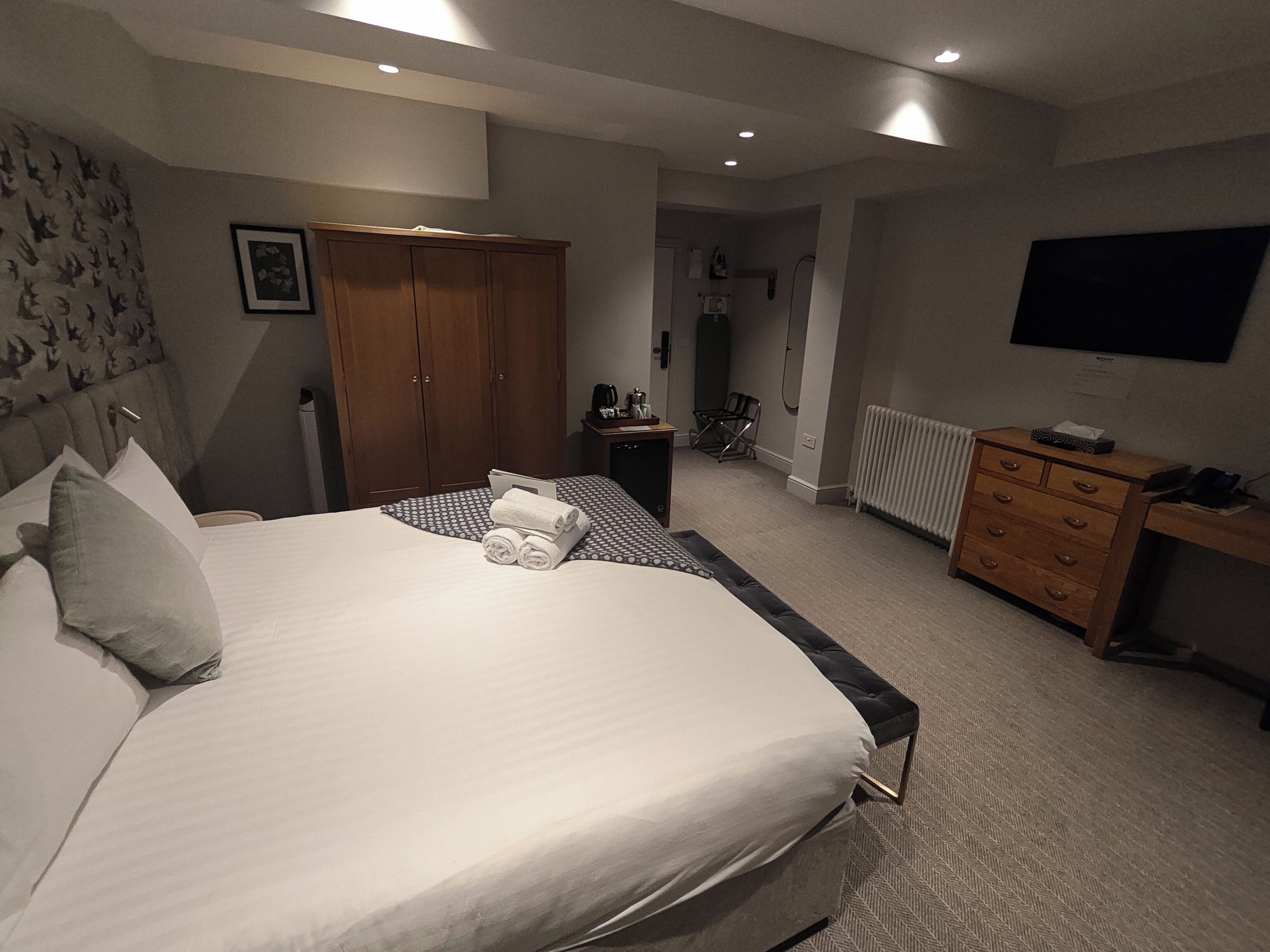 A hotel room with a large bed, a wardrobe, a desk with a chair, a radiator, and a television on the wall.