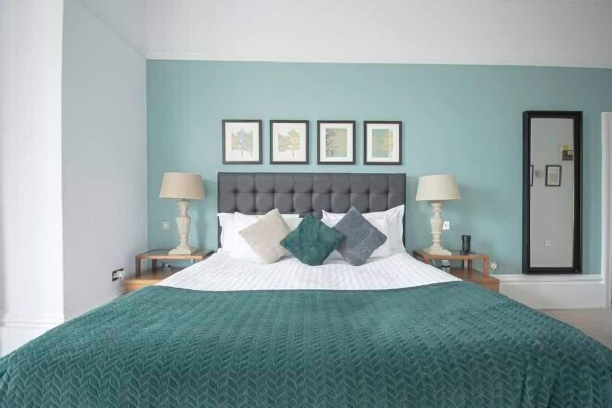 A neatly made bed with a green quilt, a grey tufted headboard, and four framed pictures on the wall.