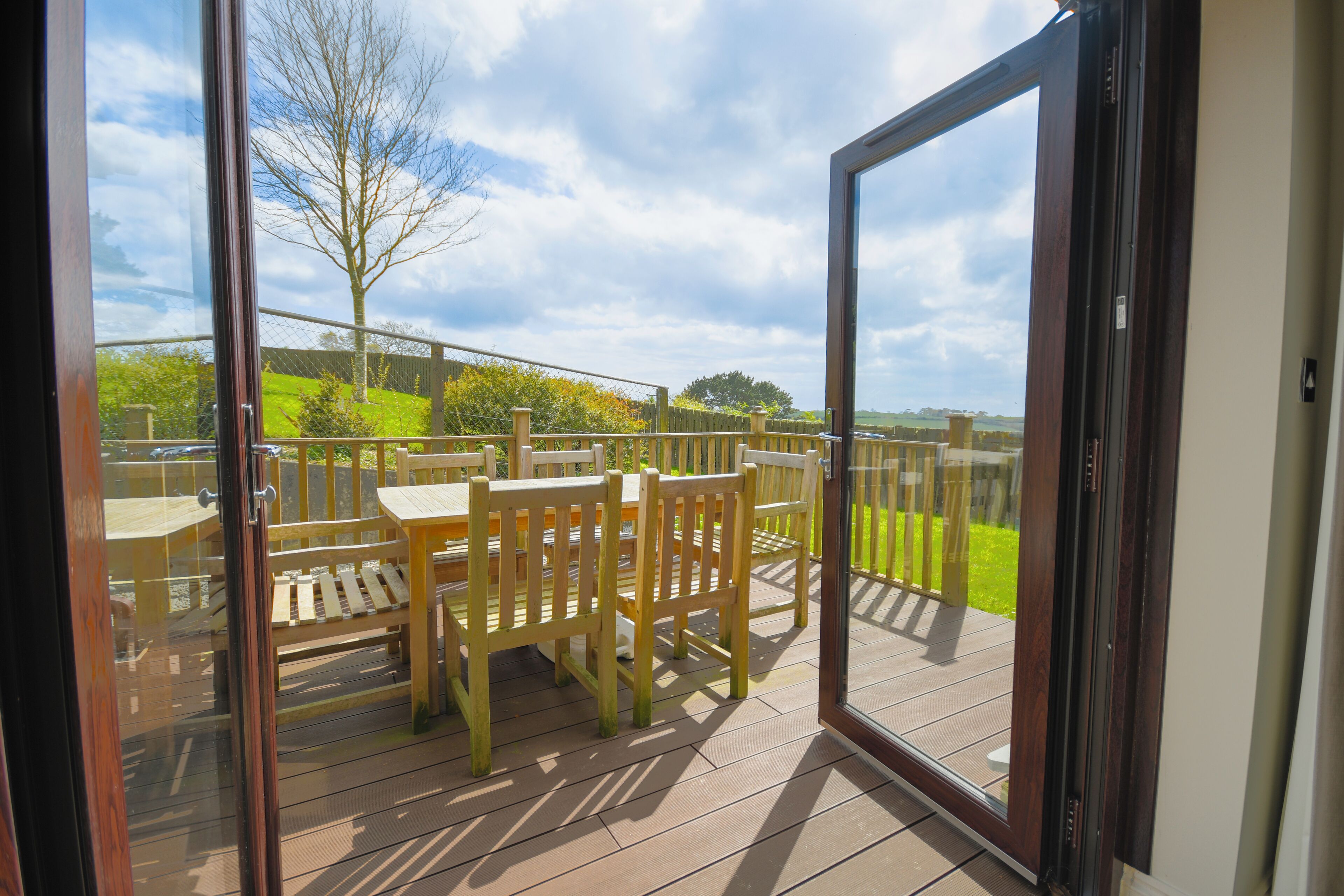 Puerta de cristal abierta que da a una terraza exterior con muebles de madera y vistas a un espacio verde.