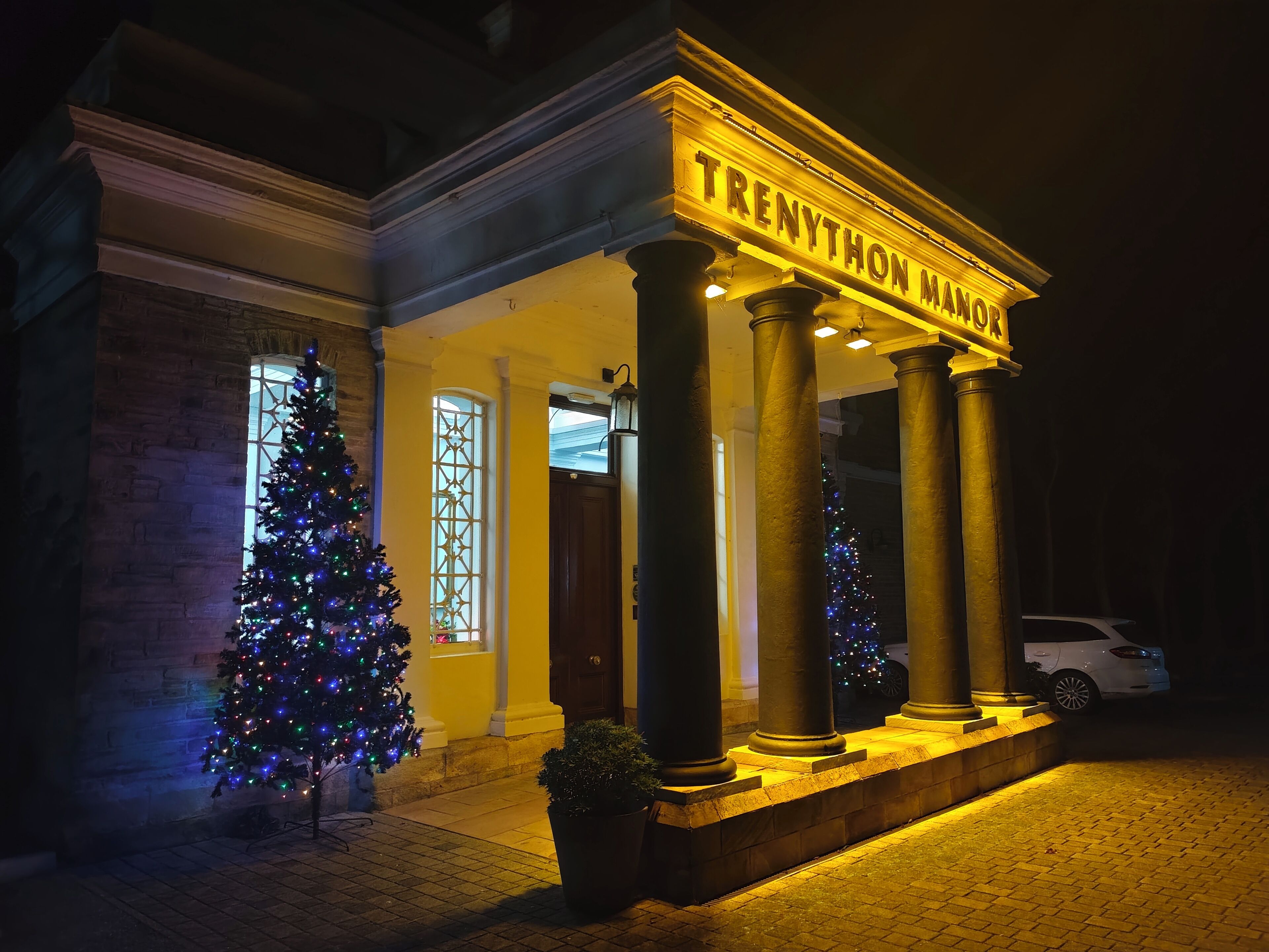 A building with "TRENYTHON MANOR" illuminated signage, large columns, and a Christmas tree in front.
