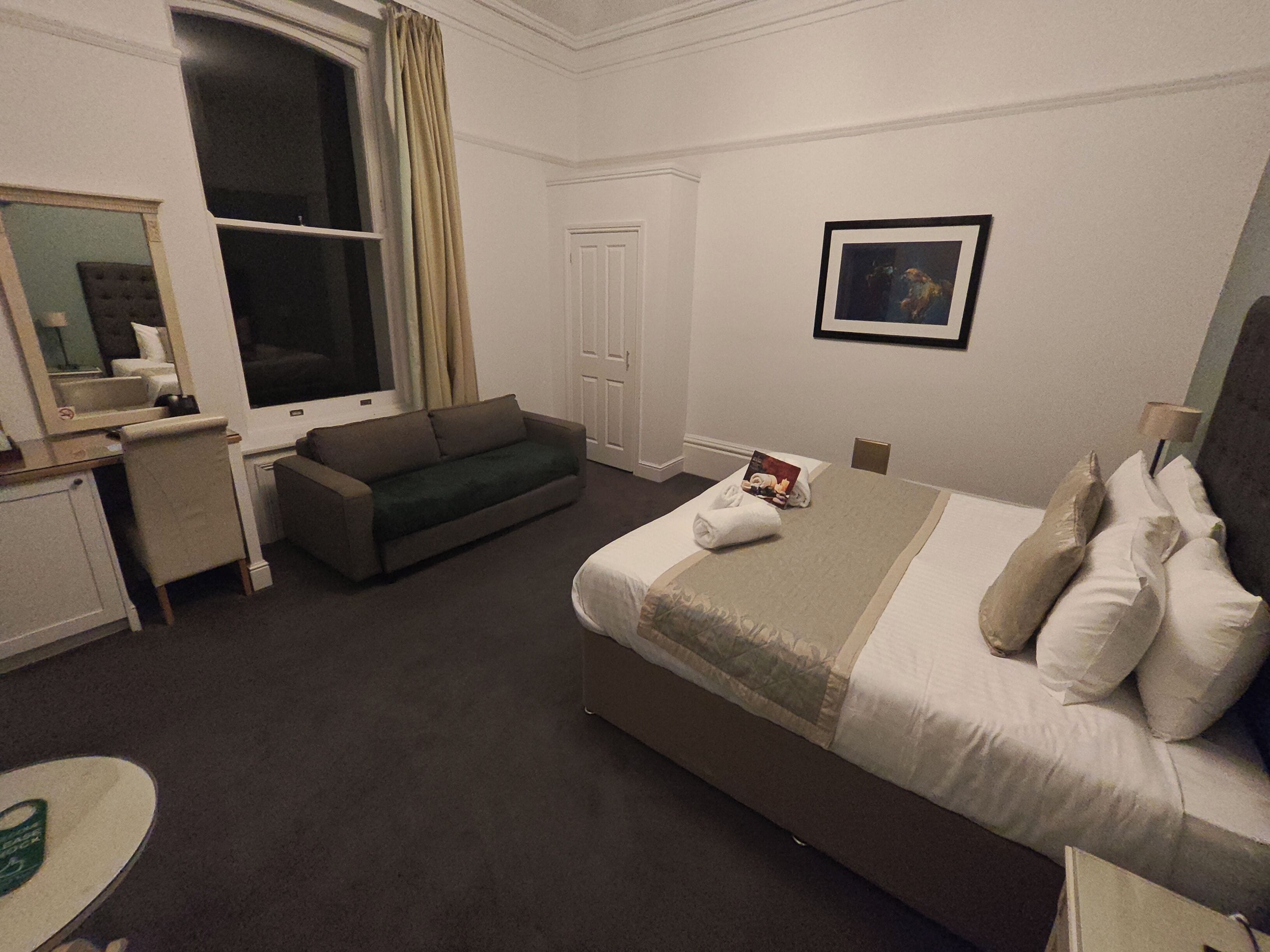 Une chambre d’hôtel comprenant un lit, un canapé, un bureau, une chaise, une fenêtre avec des rideaux, une porte et un tableau encadré au mur.