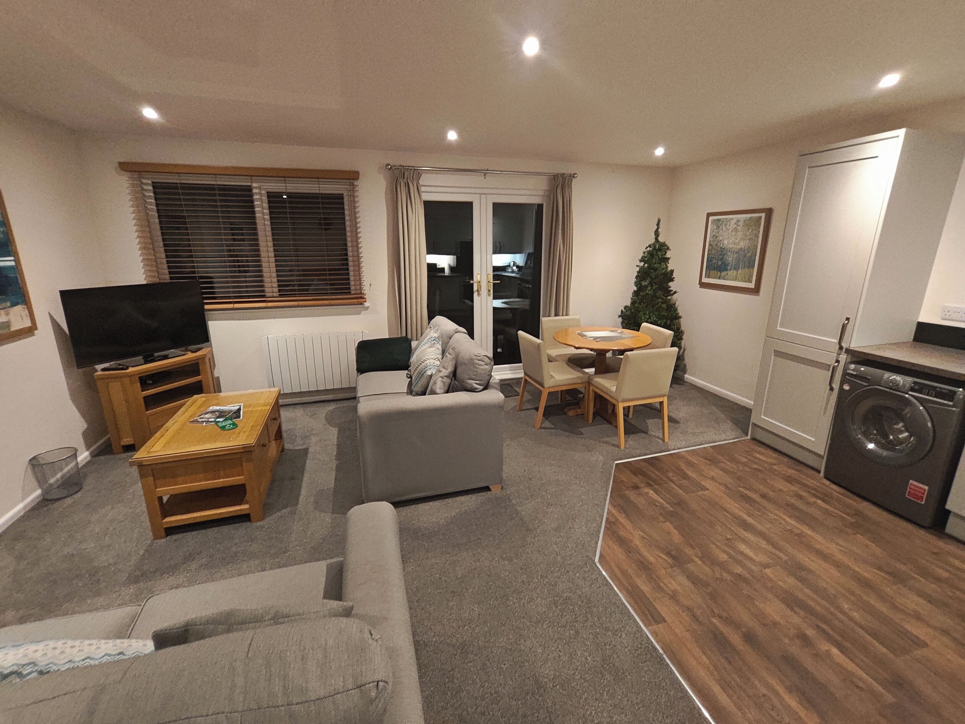 A compact living space with a sofa, a coffee table, a dining table with chairs, a small kitchen area with a washing machine, and a television on a stand.
