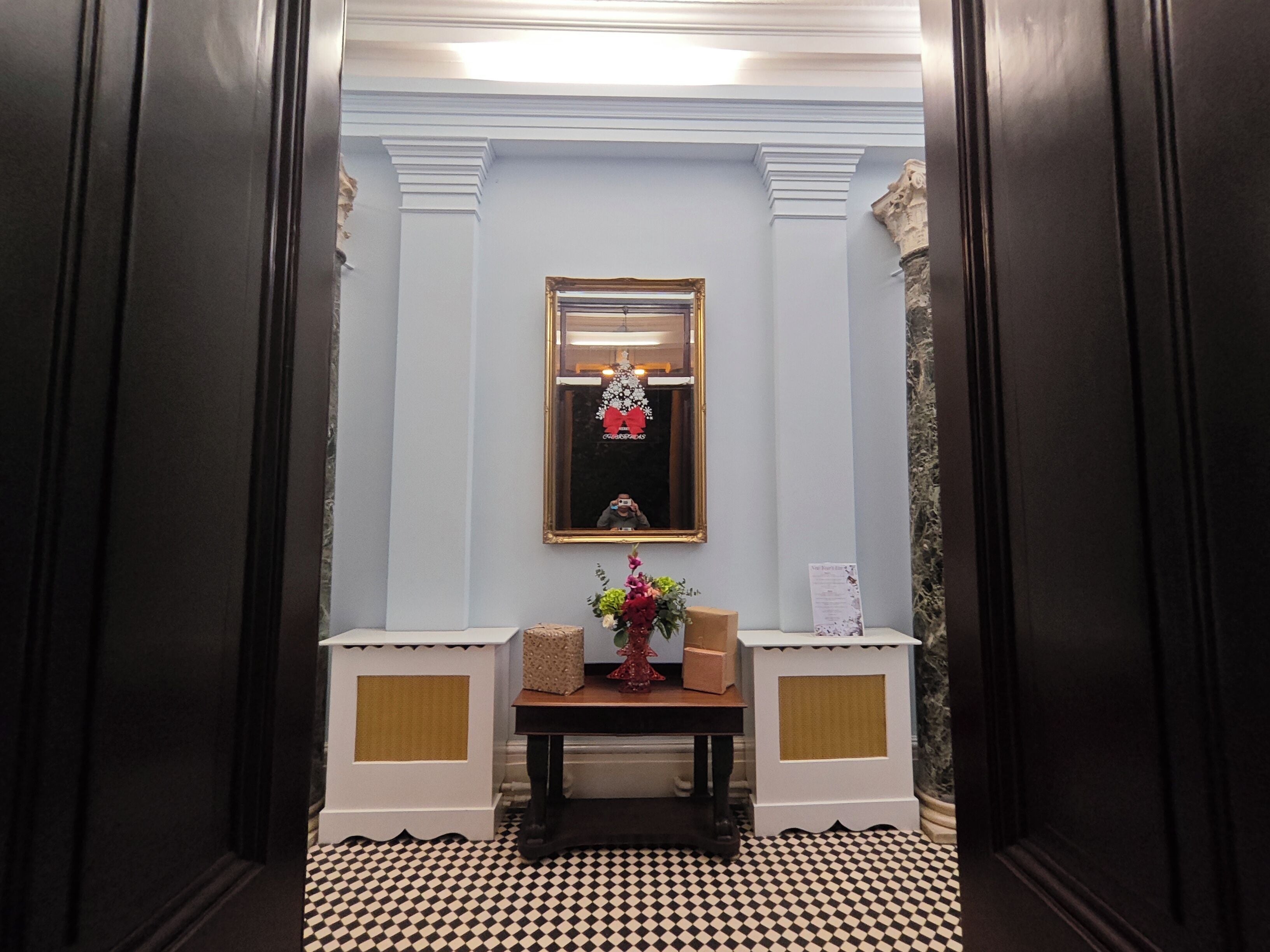 A hallway with a checkered floor, a mirror on the wall, and a small table with a vase of flowers.