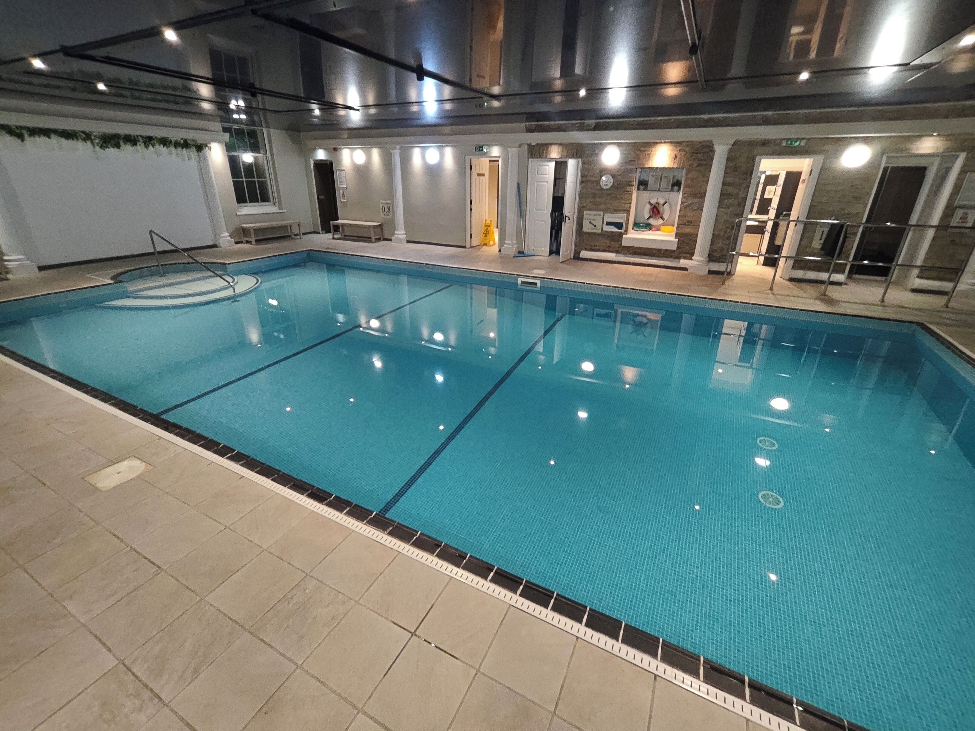 An indoor swimming pool with clear water, tiled flooring, and a bench near the pool.