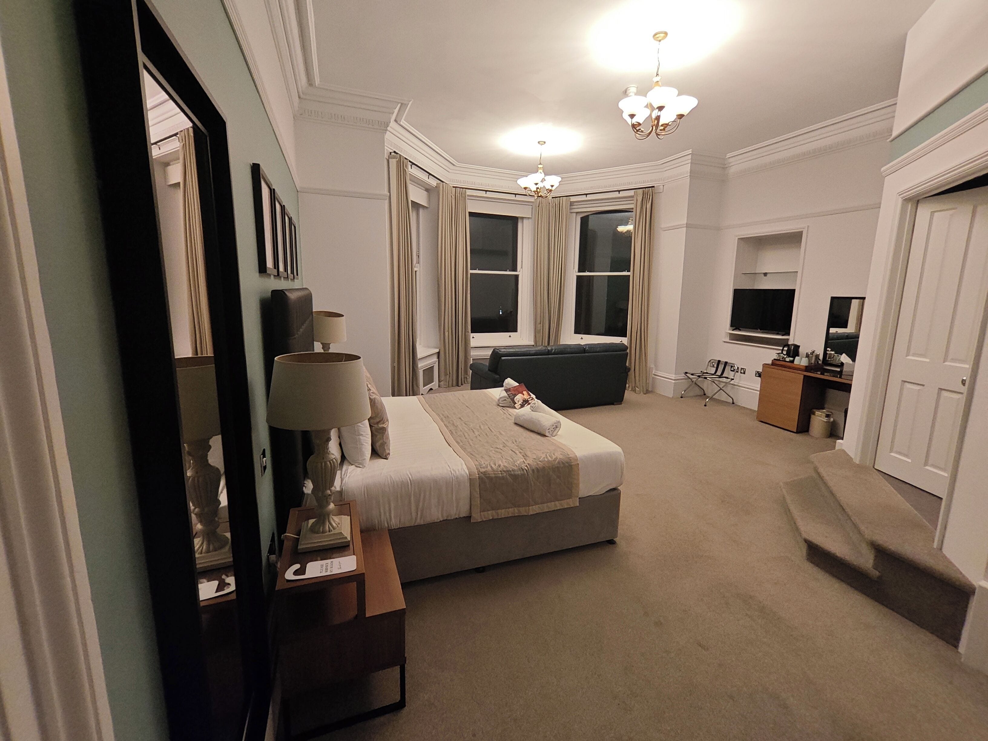A hotel room with a large bed, a desk with a lamp, a TV, and a window with curtains.