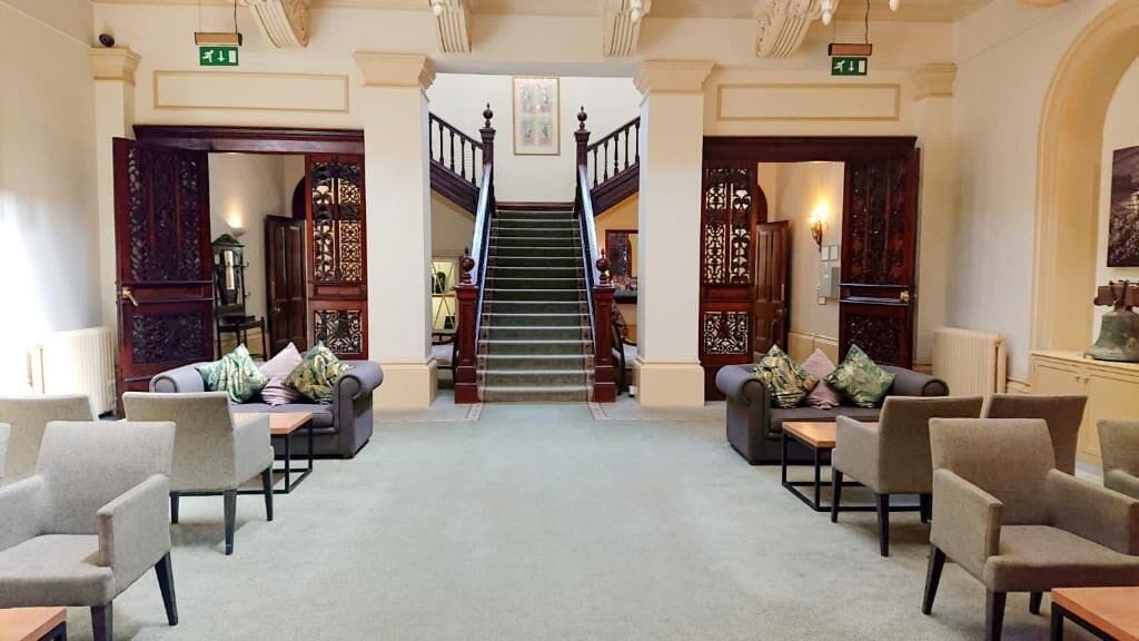 A lobby with a grand staircase, seating areas with sofas and armchairs, and traditional wooden doors.