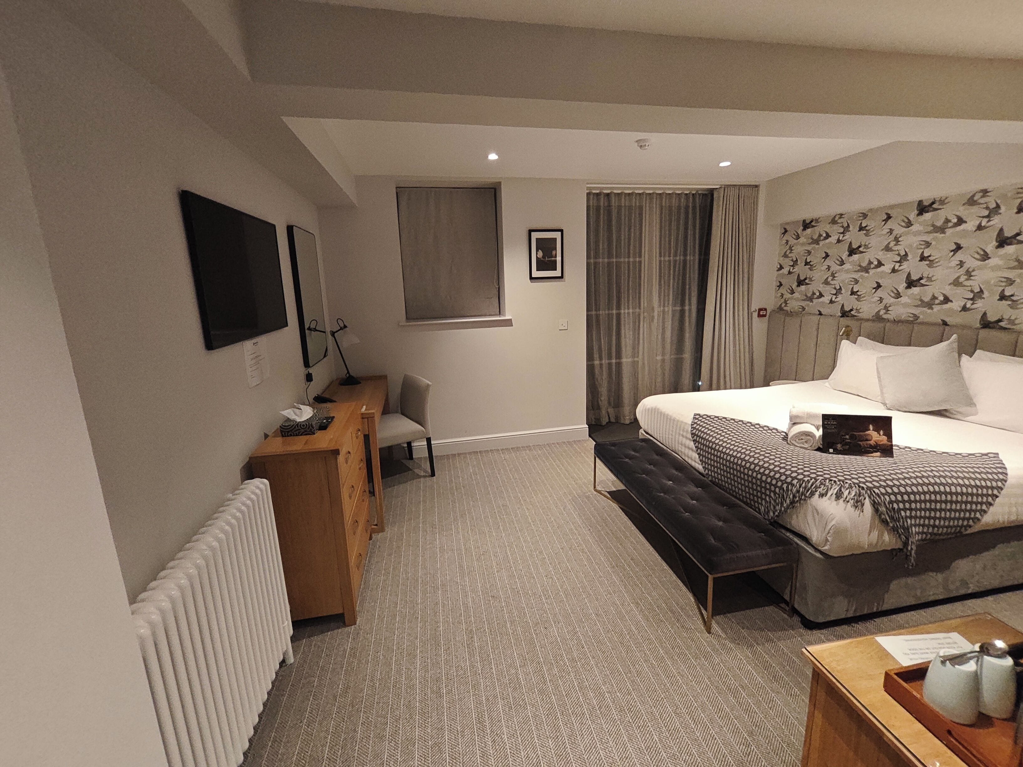 A modern hotel room with a large bed, a desk, a chair, a radiator, and a television on the wall.