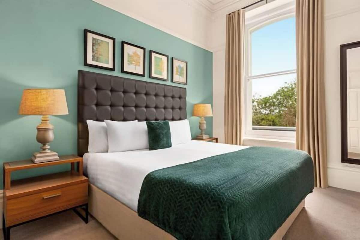 A neatly made bed with a dark headboard, two bedside lamps, and a window with curtains.