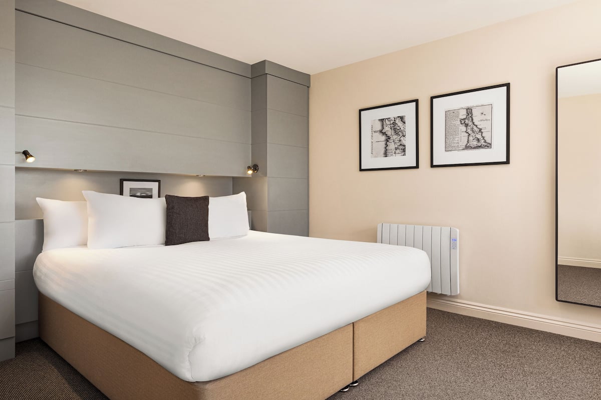 A modern hotel room with a large bed, a headboard with integrated lighting, a radiator, and two framed maps on the wall.
